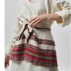 May include: Cream-colored linen dress with long sleeves and a V-neck. The waist is cinched with a decorative belt featuring a red and gold geometric pattern. The belt is tied in a bow at the front.