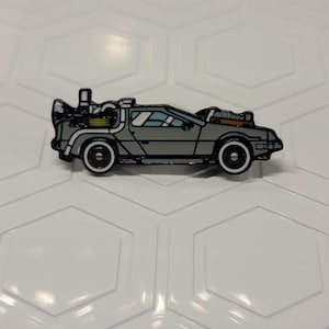 May include: A gray DeLorean car pin with black and white accents. The pin features the iconic car design with detailed elements like the flux capacitor and wheels. A collectible item for fans of the classic movie.