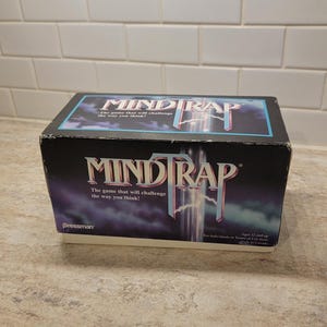 May include: A vintage Mind Trap board game box. The box is black with the game title in a stylized font, and the text "The game that will challenge the way you think!" is also visible. The game is for ages 12 and up.