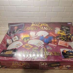 Könnte beinhalten: Ein rotbraunes Set von Lance Burton Video Magic mit 50 Tricks. Die Box zeigt Bilder von Spielkarten, Würfeln und anderen Zauberrequisiten. Das Set enthält Lehrvideos und diverses Zubehör.