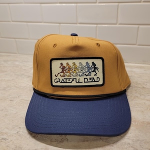 May include: Mustard yellow and navy blue baseball cap with a rectangular patch featuring a colorful graphic of dancing skeletons and the words "Grateful Dead". The cap has a curved brim and a black rope detail.
