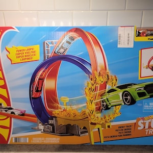 May include: Hot Wheels Stunt Tracks playset box featuring a blue and orange loop with a green car. The box has the words "Power Loops!" and "Stunt Tracks" on it. A small car is on top of the box.
