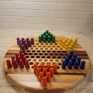 May include: A wooden Chinese checkers board with a circular design. The board features a series of holes and is surrounded by wooden stripes. Six sets of colored pegs in red, orange, yellow, green, blue, and purple are arranged on the board.