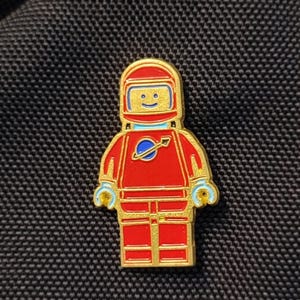 Puede incluir: Un pin de astronauta dorado con un traje espacial y casco rojos. El pin presenta una cara sonriente en el casco y un diseño de planeta azul en el pecho. El pin es una representación estilizada de una figura de Lego.
