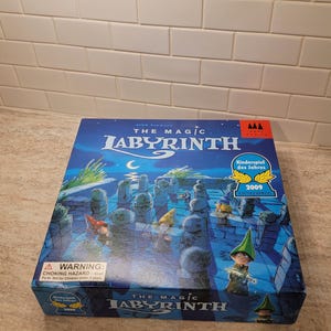 Pode incluir: Uma caixa de jogo de tabuleiro intitulada "The Magic Labyrinth" com um fundo azul e ilustrações de um labirinto e personagens de fantasia. A caixa tem um rótulo de aviso e um selo de prêmio "Kinderspiel des Jahres 2009".