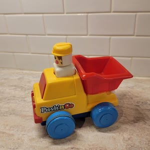 May include: A yellow and red toy dump truck with blue wheels. The truck has a small figure in the driver's seat and the words "Push'n Go" printed on the side. The toy is made of plastic.