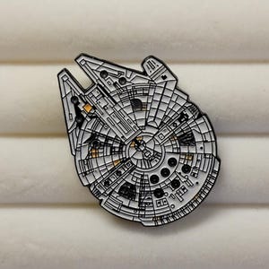 May include: A detailed enamel pin depicting the Millennium Falcon starship from Star Wars. The pin features a white and black design with orange accents, showcasing the ship's intricate details. This collectible item is perfect for fans of science fiction.