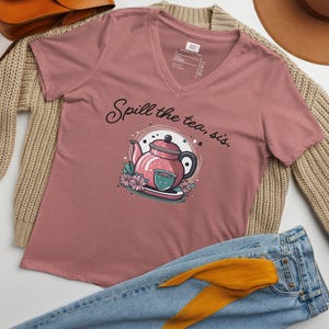 May include: A mauve v-neck t-shirt with the text "Spill the tea, sis." and a graphic of a pink teapot with a teacup and flowers.