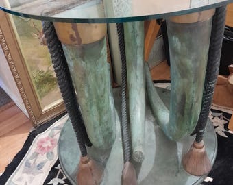 May include: A glass-topped table with a unique base made of three green, curved, and textured supports. The supports are connected by black rope-like accents and have wooden feet.