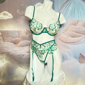 May include: A cream-coloured lingerie set with green floral embroidery. The set includes a bra, garter belt, and matching underwear. The bra has adjustable straps and a gold-coloured clasp. The garter belt has attached straps with green tassels.