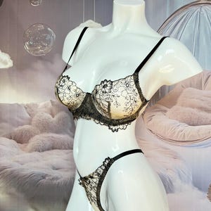 May include: Cream-coloured bra and thong set with black floral lace detailing. The lingerie is displayed on a white mannequin. The bra has adjustable straps and underwire support. The thong features a delicate lace trim.