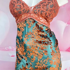 May include: A teal slip dress with a floral pattern in shades of orange and brown. The bodice features a lace overlay in a coral color. The dress has thin straps and a V-neckline.