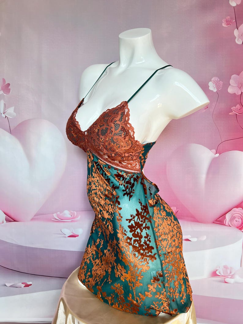 May include: A teal and rust-colored slip dress on a mannequin. The bodice features a rust-colored lace design, with thin green straps. The dress has a floral pattern and a flowing skirt. The mannequin is on a gold-colored surface.