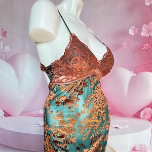 May include: A teal and brown floral patterned slip dress on a mannequin. The dress features a brown lace bodice with thin straps and a teal skirt with a brown floral design. The mannequin is white and the background is pink.