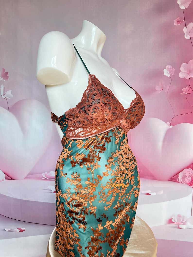 May include: A teal and brown floral patterned slip dress on a mannequin. The dress features a brown lace bodice with thin straps and a teal skirt with a brown floral design. The mannequin is white and the background is pink.