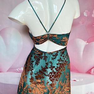 May include: A teal-colored slip dress with a floral pattern in a darker shade of brown. The dress features thin straps and a low back. The floral pattern is a velvet-like material. The dress is displayed on a white mannequin.