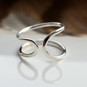 Puede incluir: Un anillo de plata con un diseño único, abierto y ondulado. El anillo está hecho de una sola pieza de metal que se dobla en un bucle.