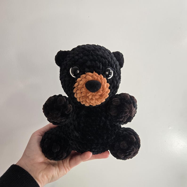 May include: A handmade black bear plush toy with a tan muzzle and large, round eyes. The bear is made of a soft, textured material and has small, rounded ears and paws. The bear is approximately 8 inches tall.