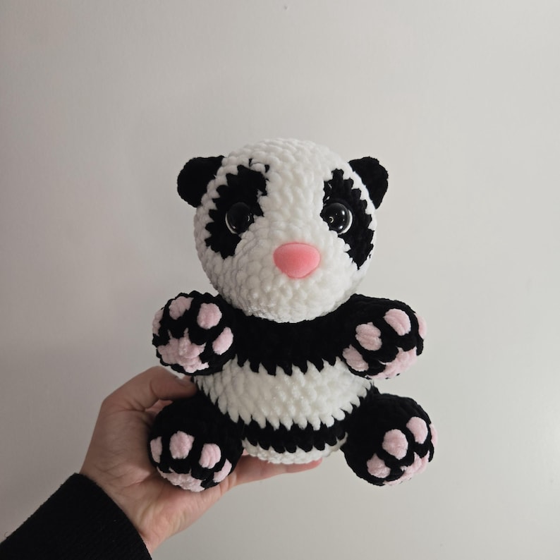 May include: A handmade plush panda bear with a white body, black markings, and a pink nose. The panda has large, expressive eyes and pink accents on its paws. This soft toy is perfect for children and panda enthusiasts.