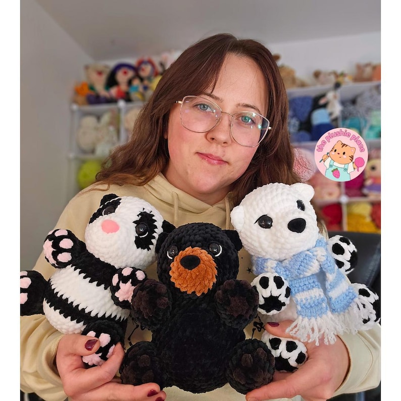 May include: Three handmade crochet plush bears: a panda, a black bear, and a polar bear. The panda has black and white fur with pink accents. The black bear has a brown snout. The polar bear wears a blue and white striped scarf.