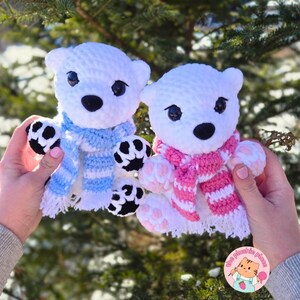 May include: Two white crochet polar bear plushies with black eyes and noses. One bear wears a blue and white striped scarf, the other a pink and white striped scarf. The bears have black and white paws and are held up against a green background.