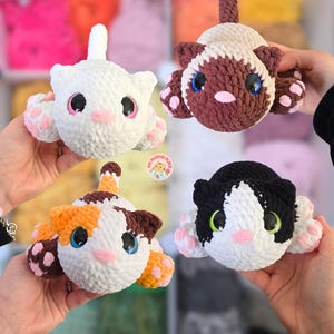May include: Four handmade crochet cat plushies in white, brown, calico, and black and white. Each cat has large, colorful eyes, pink noses, and paw pads. The plushies are held up by hands, showcasing their soft, cuddly appearance.