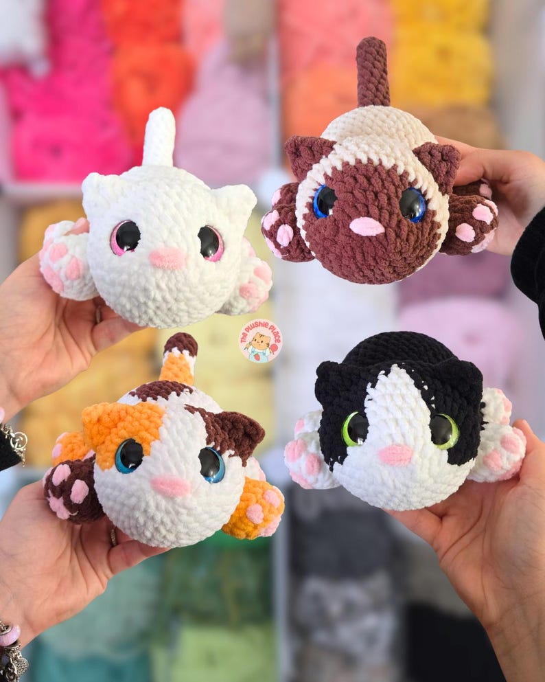 May include: Four handmade crochet cat plushies in white, brown, calico, and black and white. Each cat has large, colorful eyes, pink noses, and paw pads. The plushies are held up by hands, showcasing their soft, cuddly appearance.