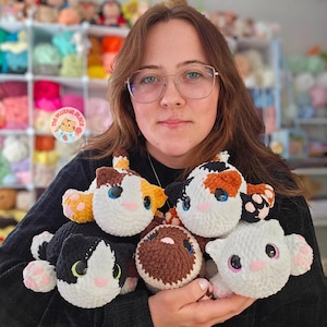 May include: A collection of handmade crochet cat plushies in various colors, including calico, black and white, and brown. Each plushie features detailed facial features and pink accents. The plushies are soft and cuddly, perfect for cat lovers.