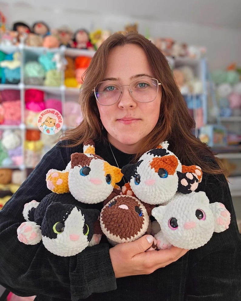 May include: A collection of handmade crochet cat plushies in various colors, including calico, black and white, and brown. Each plushie features detailed facial features and pink accents. The plushies are soft and cuddly, perfect for cat lovers.