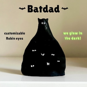 Może przedstawiać: Czarna figurka Batdada z świecącymi białymi oczami. Tekst "Batdad" znajduje się nad figurką, z "customizable Robin eyes" i "we glow in the dark!" po bokach. Unikalna dekoracja na Halloween.