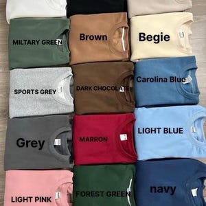K&ouml;nnte beinhalten: Eine Auswahl an Sweatshirts mit Rundhalsausschnitt in verschiedenen Farben, darunter Wei&szlig;, Schwarz, Sand, Milit&auml;rgr&uuml;n, Braun, Beige, Sportgrau, Dunkelbraun, Grau, Kastanienbraun, Hellblau, Hellrosa, Waldgr&uuml;n und Marineblau. Jedes Sweatshirt hat ein Etikett mit seinem Farbnamen.