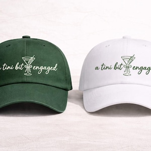 Puede incluir: Se muestran dos gorras de béisbol, una verde oscuro y otra blanca. Cada gorra tiene la frase bordada "a tini bit engaged" y un diseño de copa de Martini a juego. Accesorios de cabeza informales.