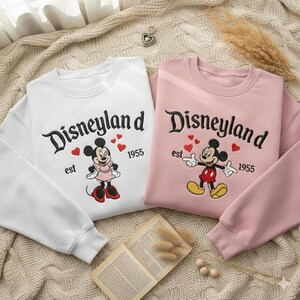 Może przedstawiać: Dwie bluzy, jedna biała i jedna r&oacute;żowa, z logo Disneylandu oraz Myszki Miki i Myszki Minnie. Biała bluza ma Myszkę Minnie, a r&oacute;żowa Myszkę Miki. Obie mają napis "est 1955".