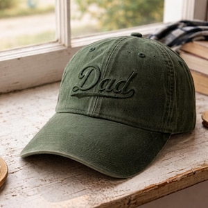 Op de afbeelding: Olijfgroene baseballcap met het woord "Dad" in cursief geborduurd. De cap heeft een gebogen klep en een klassiek zes-panelen ontwerp. De hoed is gemaakt van een zacht, versleten materiaal.