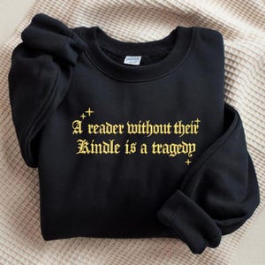 May include: Black sweatshirt with gold text that reads "A reader without their Kindle is a tragedy." The sweatshirt is folded on a beige, textured surface. The text is in a gothic font.