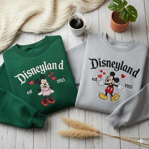 Może przedstawiać: Dwie bluzy, jedna zielona i jedna szara, z logo Disneylandu i grafiką Myszki Miki i Myszki Minnie. Zielona bluza przedstawia Myszkę Minnie, a szara Myszkę Miki. Obie zawierają napis "est 1955".