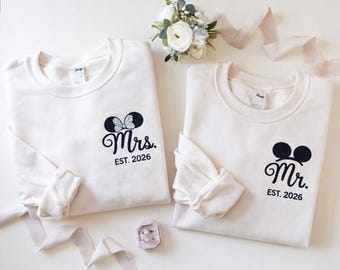 May include: Two off-white sweatshirts with black Mickey Mouse ear designs and the text "Mrs. EST. 2026" and "Mr. EST. 2026". A small bouquet of white flowers and a ring box are also present.