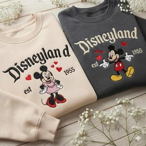 Może przedstawiać: Dwie bluzy z okrągłym dekoltem, jedna kremowa i jedna szara, z napisem "Disneyland" w ozdobnym kroju pisma. Kremowa bluza ma grafikę Minnie Mouse, a szara Myszki Miki. Obie mają rok "1955" i czerwone serca.