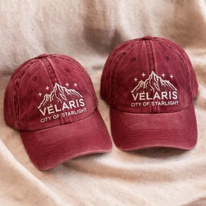 May include: Two maroon baseball caps with the text "Vēlaris City of Starlight" in white. The caps feature a mountain and star design. The caps are made of a soft, worn material.