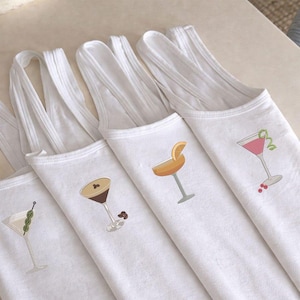 May include: White tank tops with embroidered cocktail designs. Each top features a different cocktail illustration: a martini with olives, an espresso martini, a margarita with an orange slice, and a pink cocktail with a garnish.