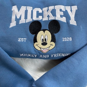 K&ouml;nnte beinhalten: Hellblaues Sweatshirt mit wei&szlig; gesticktem Schriftzug "MICKEY". Darunter ein l&auml;chelndes Mickey Mouse-Gesicht, mit "EST 1928" und "MICKEY AND FRIENDS" ebenfalls gestickt. Das Sweatshirt hat gerippte B&uuml;ndchen und einen Rundhalsausschnitt.