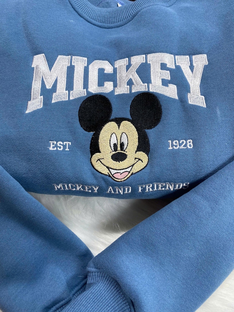 K&ouml;nnte beinhalten: Hellblaues Sweatshirt mit wei&szlig; gesticktem Schriftzug "MICKEY". Darunter ein l&auml;chelndes Mickey Mouse-Gesicht, mit "EST 1928" und "MICKEY AND FRIENDS" ebenfalls gestickt. Das Sweatshirt hat gerippte B&uuml;ndchen und einen Rundhalsausschnitt.