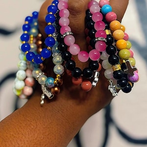 May include: A collection of colorful beaded bracelets with various charms, including a cross and a bird. The bracelets are made with different types of beads, including pink, blue, black, and green.
