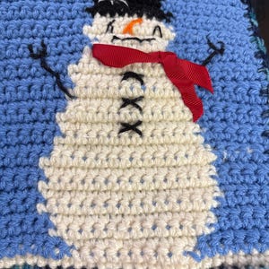 May include: Crocheted snowman design on a blue background. The snowman has a white body, black hat, orange nose, and a red ribbon scarf. The crochet work is detailed, with black stitches for the eyes and mouth.
