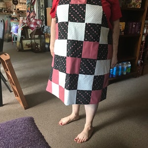 May include: A patchwork apron with a checkerboard design in black, white, and pink. The apron features a floral pattern on some squares and a solid pink on others. It has white straps and is worn by a person.