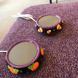 May include: Two purple and black Halloween-themed electric wax warmers with a round, flat surface. One warmer has a candy corn design and the other has a pumpkin design.