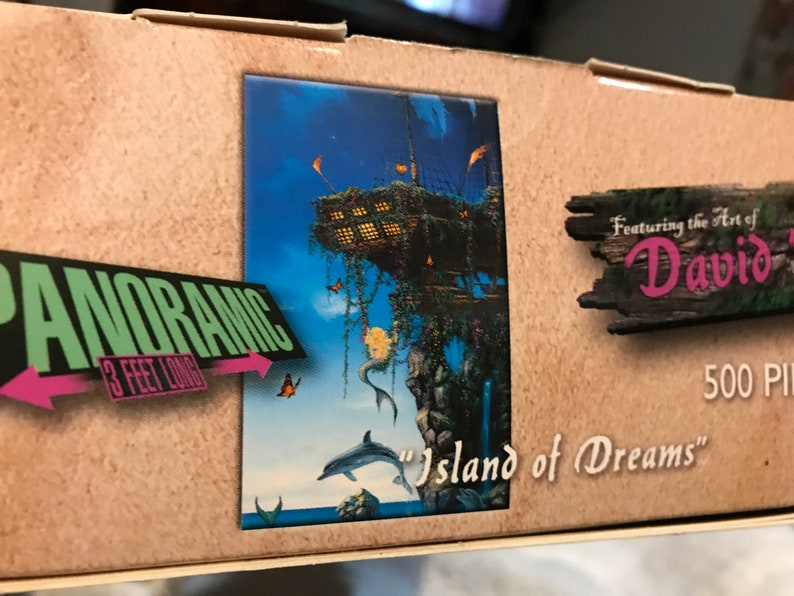May include: A 500 piece panoramic jigsaw puzzle featuring the art of David. The puzzle depicts a whimsical island with a treehouse, a mermaid, and a dolphin. The box is brown with a pink and green "Panoramic 3 feet long" label.
