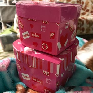 May include: Two stacked pink gift boxes with heart and floral patterns. The boxes have the words "kisses" and "my love" printed on them. A ribbon is tied on top of the boxes.