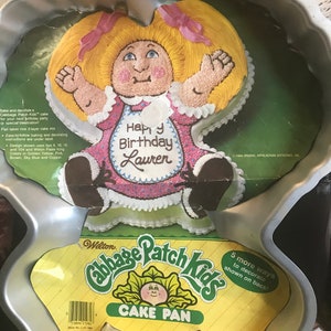 May include: A silver metal cake pan shaped like a Cabbage Patch Kid doll. The pan is designed to make a two-layer cake. The pan has a yellow label that says "Wilton Cabbage Patch Kids Cake Pan". The label also has a picture of a Cabbage Patch Kid doll with the text "Happy Birthday Lauren".