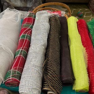 May include: A collection of eight rolls of decorative mesh fabric in various colors and textures, including white, red and green plaid, silver, black with gold accents, solid black, neon yellow, solid red, and solid green, all arranged side by side.